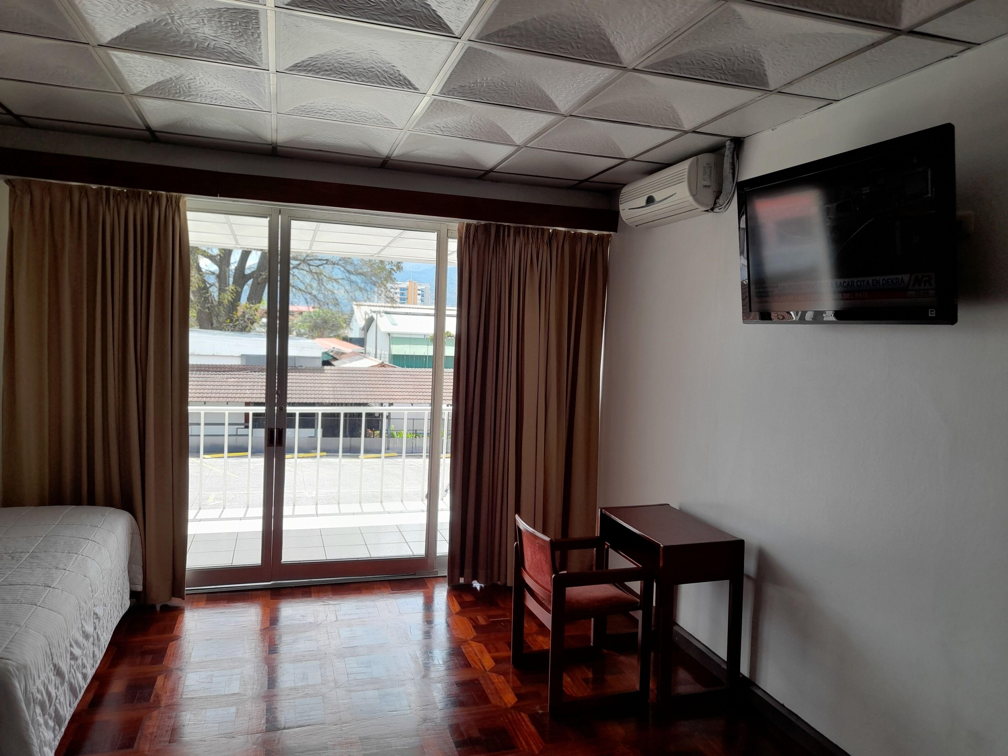 Une chambre d’hôtel équipée d’un lit, d’un bureau, d’une chaise, d’une télévision et d’une porte-fenêtre coulissante donnant sur l’extérieur.
