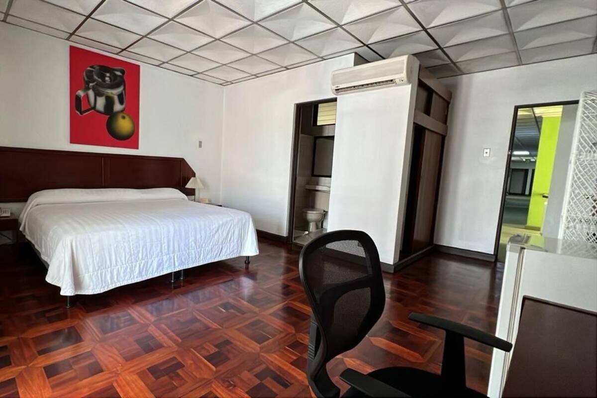 A hotel room with a wooden parquet floor, a bed with a white bedspread, a desk with a chair, a red artwork on the wall, and an air conditioning unit.