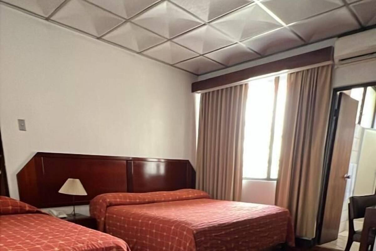A hotel room with two beds, wooden flooring, a large window with curtains, and a ceiling with a geometric pattern.