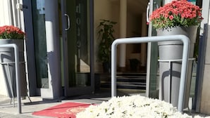 Entrada de un hotel con puerta de vidrio, un felpudo rojo y dos macetas con flores rojas.
