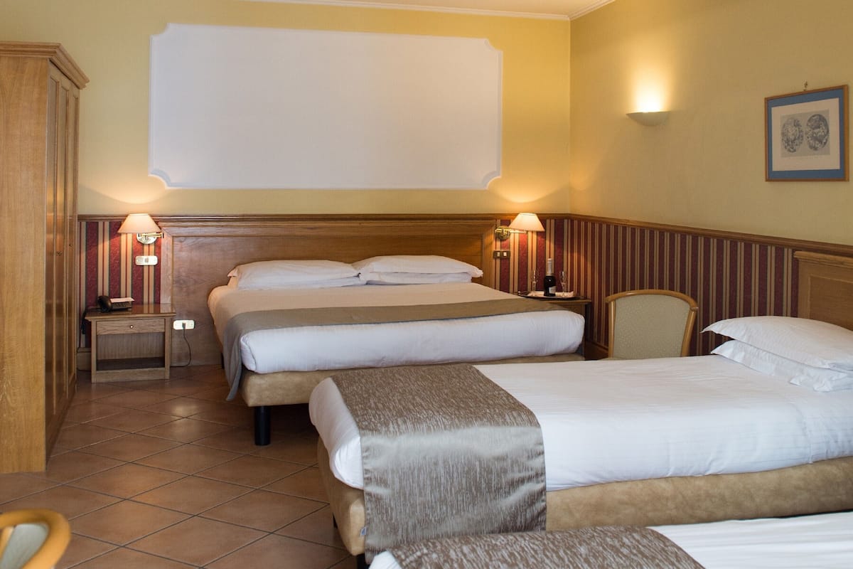 A hotel room with two beds, a wooden wardrobe, a nightstand with a lamp, and a framed picture on the wall.