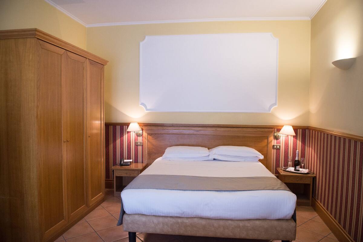 A hotel room with a large bed, wooden wardrobe, two bedside tables with lamps, and a blank wall above the bed.