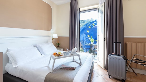 Habitación de hotel con cama, bandeja de desayuno, maleta y vista de la bandera de la Unión Europea fuera.