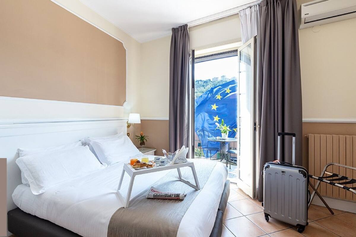 A hotel room with a bed, a tray with breakfast items, a suitcase, and a view of the European Union flag outside.
