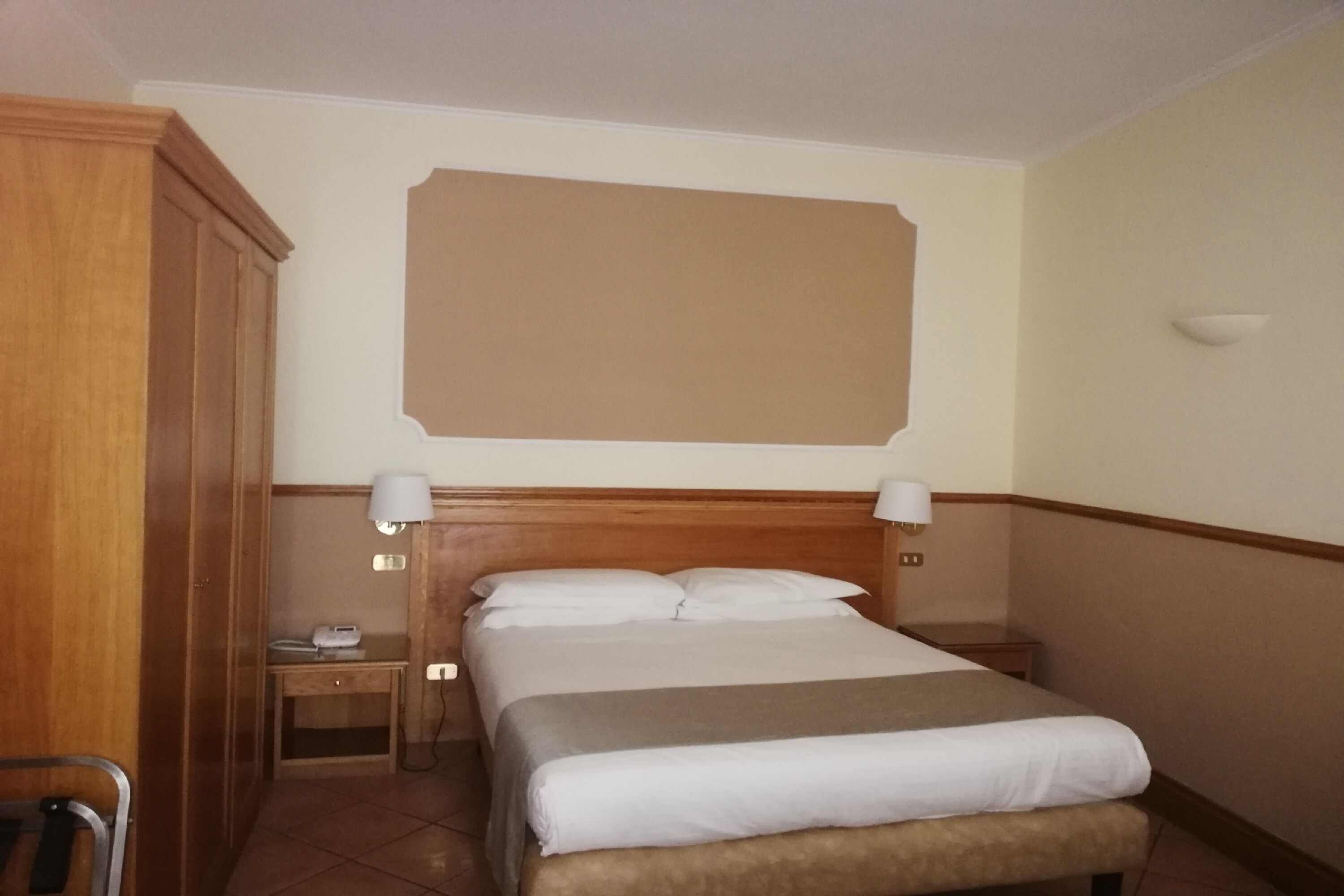 Une chambre d’hôtel avec une tête de lit en bois, un lit avec une literie blanche, deux lampes de chevet, une table de chevet et une chaise.