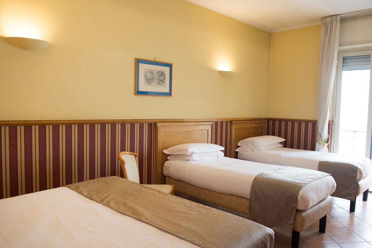 A hotel room with three single beds, a wooden headboard, striped wallpaper, a framed picture on the wall, and a window with curtains.