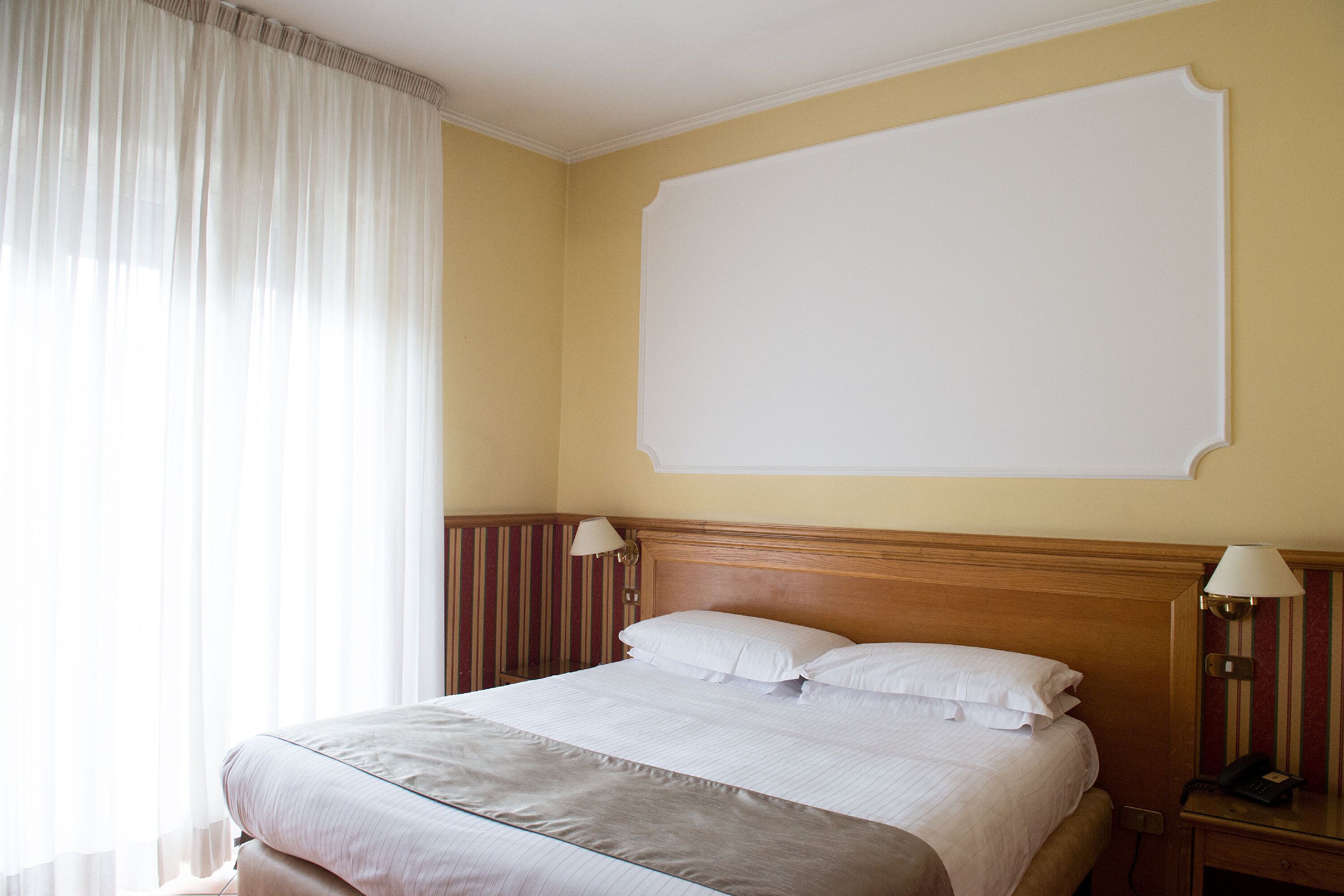 A hotel room with a large bed, two bedside lamps, a plain wall, and a window with sheer curtains.