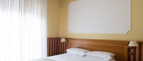 Habitación de hotel con una cama grande, dos lámparas de noche, una pared lisa y una ventana con cortinas translúcidas.