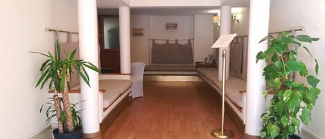 Un pasillo de hotel con suelo de madera, paredes blancas y columnas. Se observan dos plantas en macetas, una cama y un escritorio con una lámpara.