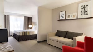 A hotel room with a bed, a sofa, a red armchair, a desk, and a lamp. There are framed pictures on the wall.