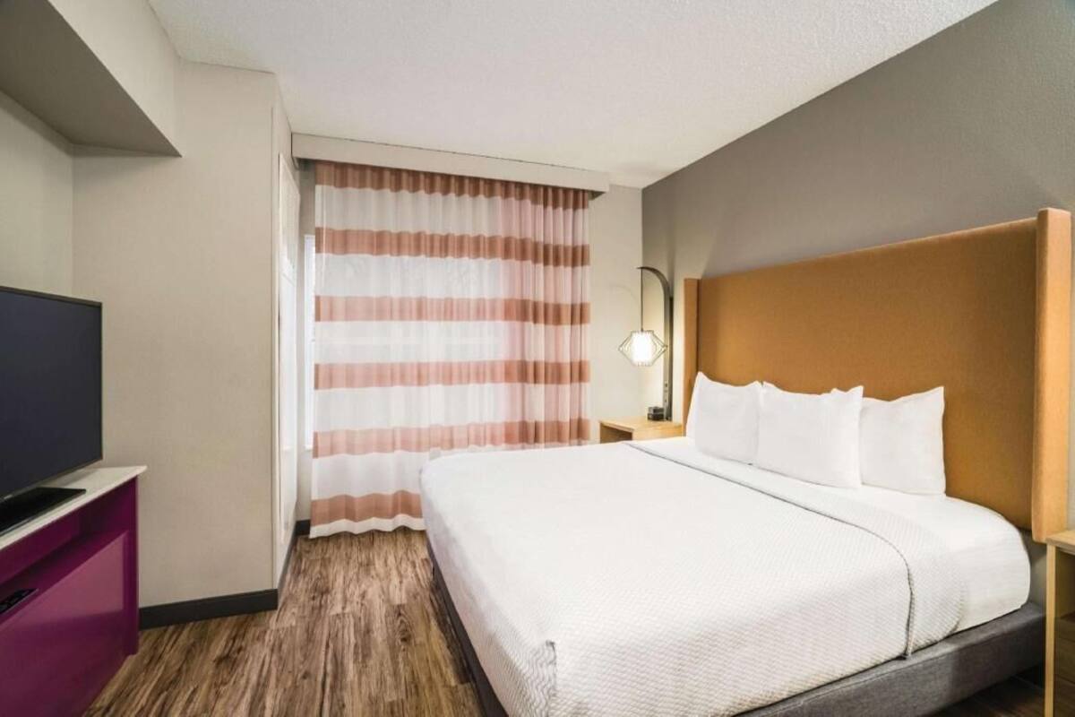 Ein Hotelzimmer mit Bett, Fernseher, Nachttisch mit Lampe und einem Fenster mit Vorhängen.