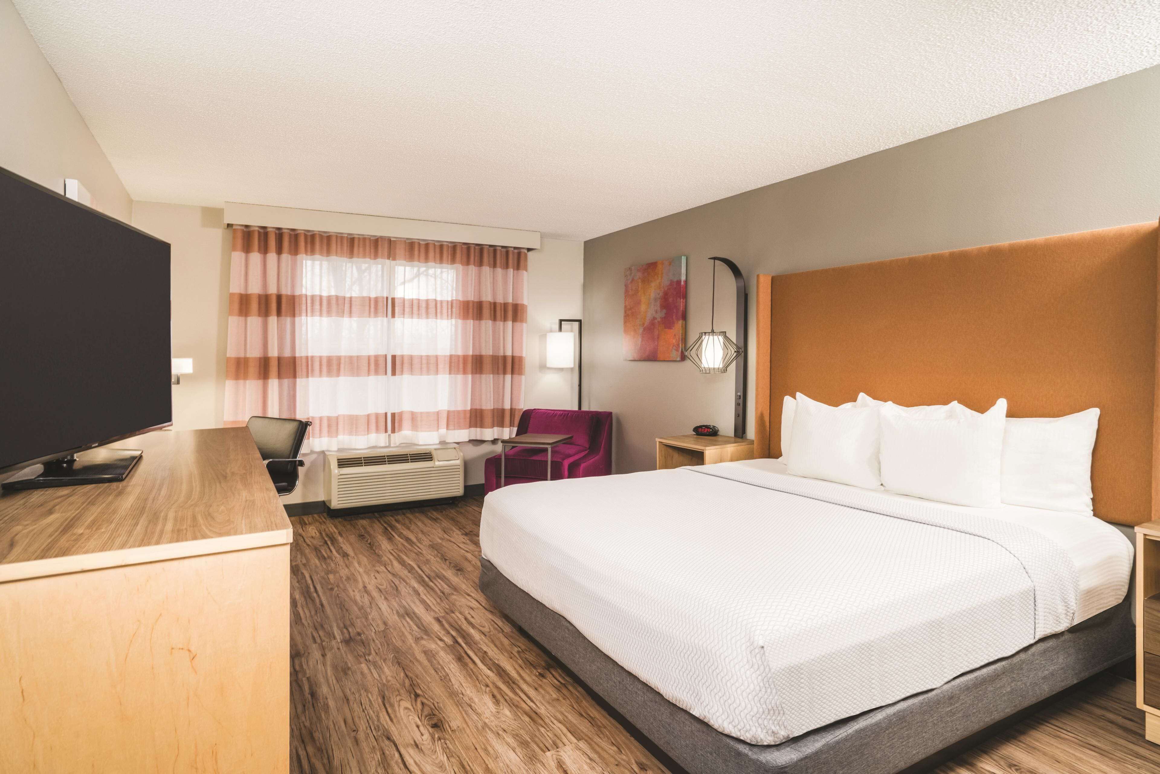 A hotel room with a large bed, a desk with a flat-screen TV, a chair, a desk lamp, and a wall-mounted artwork.