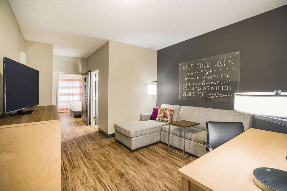 Ein modernes Wohnzimmer mit einer Eckcouch, einem Flachbildfernseher, einem Holztisch und einer Tafelwand mit motivierenden Sprüchen.