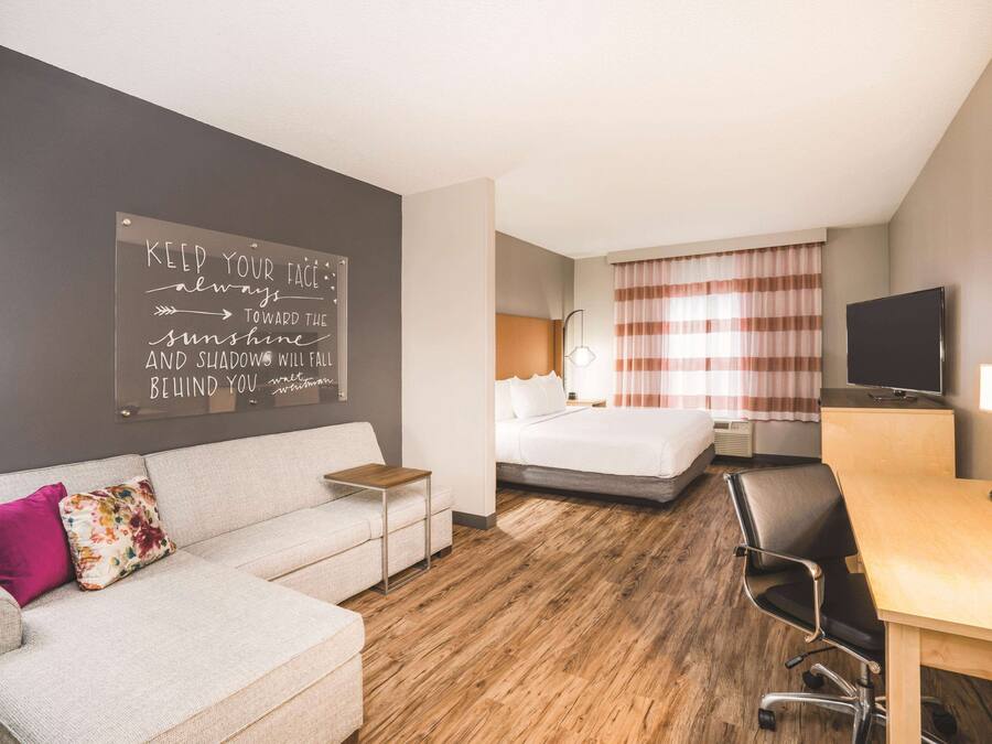 Une chambre d’hôtel équipée d’un lit, d’un bureau, d’une chaise, d’une télévision et ornée d’une citation inspirante sur le mur.