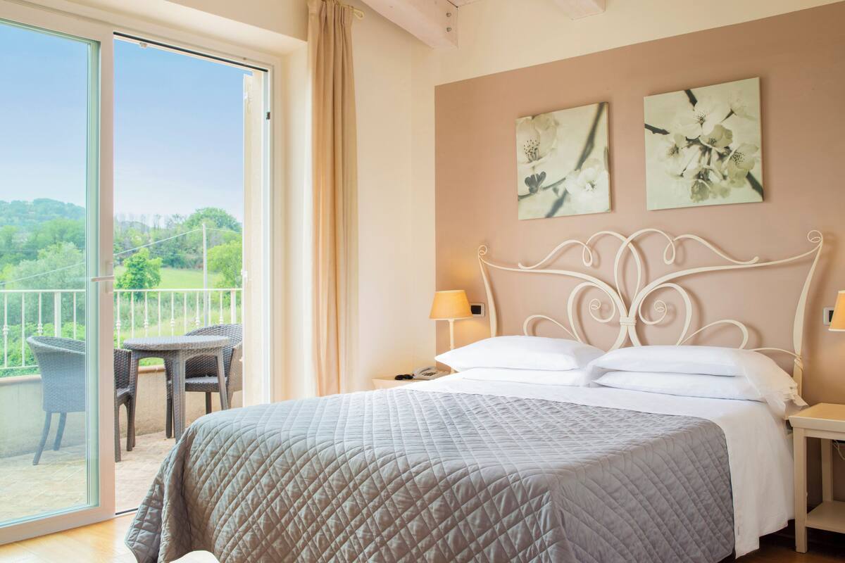 A bedroom with a bed, a nightstand, a lamp, a chair, and a balcony with a view of greenery.