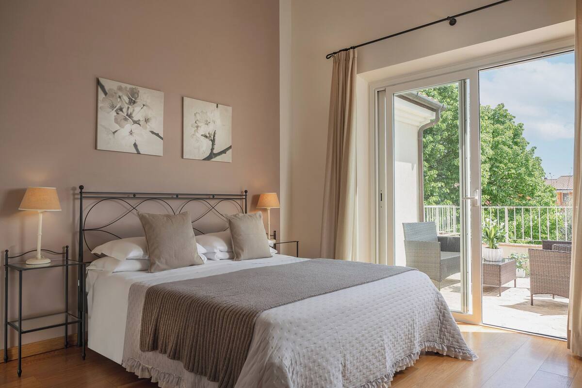 A bedroom with a bed, two bedside tables, a window with curtains, and two wall-mounted artworks.
