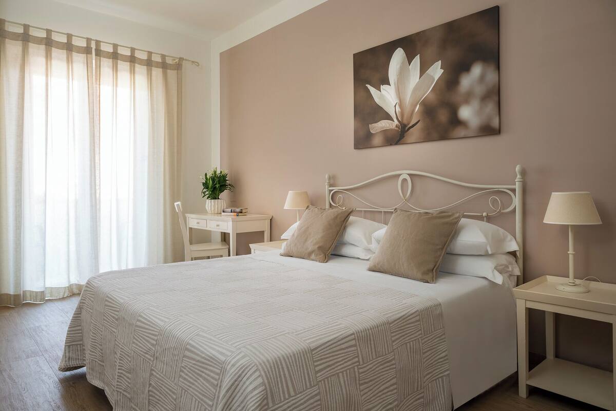 A bedroom with a large bed, two bedside tables, a nightstand, a desk with a chair, and a window with curtains.