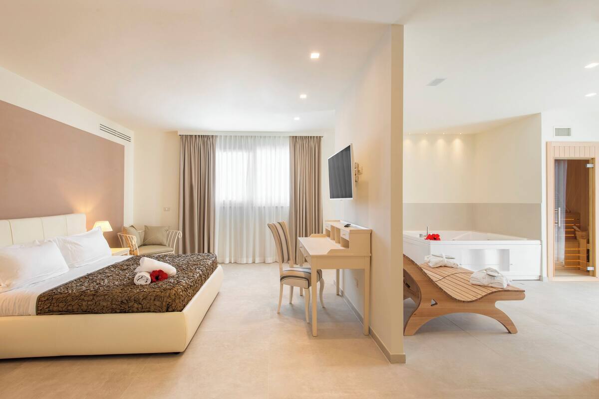 A modern hotel room with a large bed, a desk, a chair, a wooden bench, a TV, and a bathroom with a bathtub.