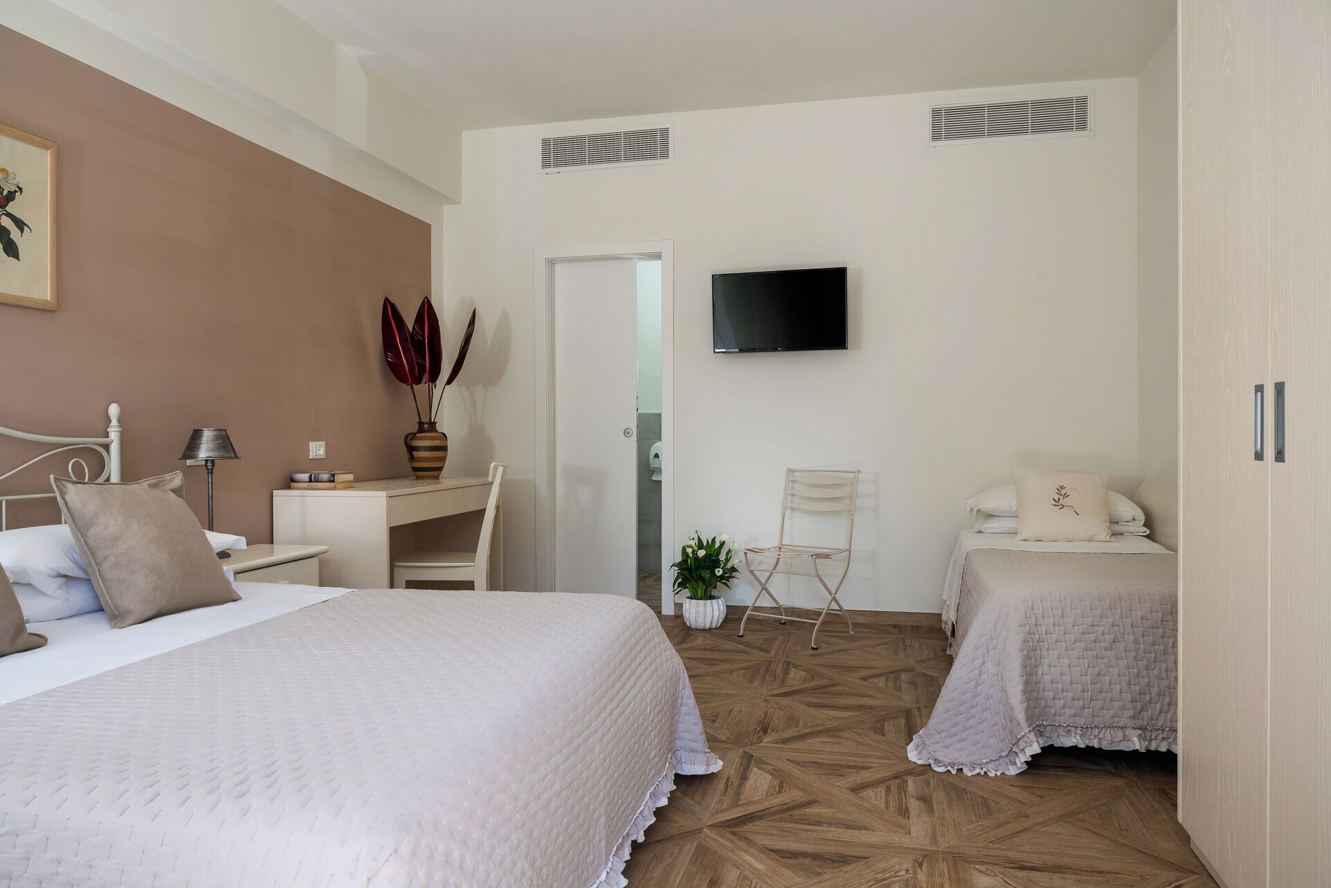 A hotel room with two beds, a desk, a chair, a TV, and a potted plant.