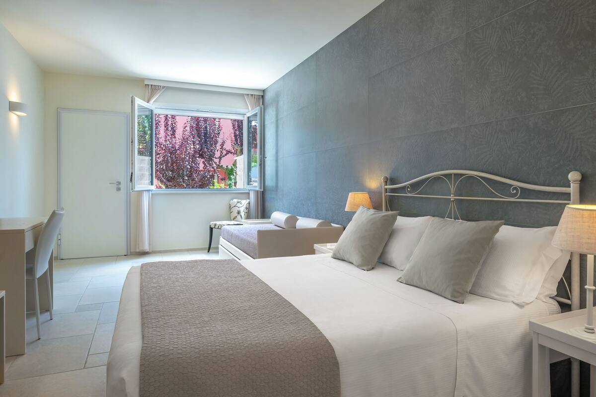 A modern bedroom with a large bed, bedside lamps, a window with a view of foliage, and a door.