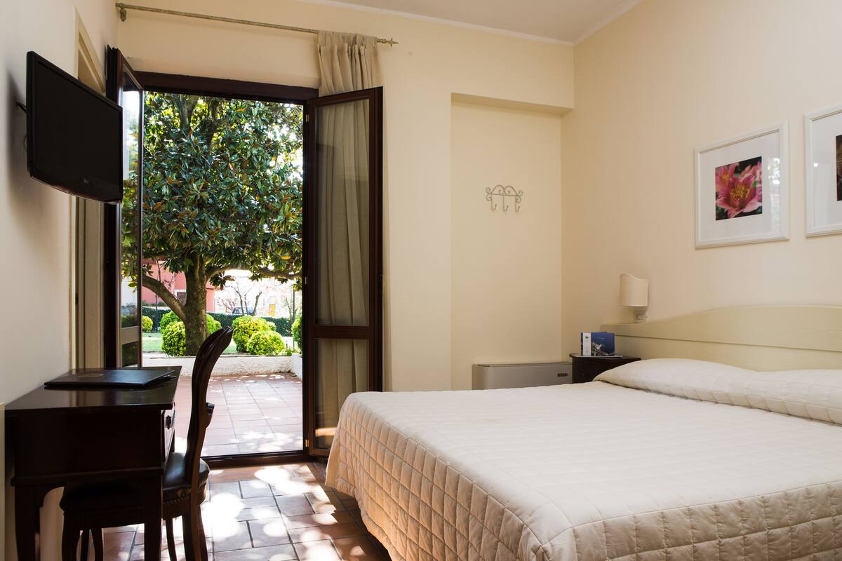 A hotel room with a bed, a desk, a chair, a TV, and a window with a view of greenery.
