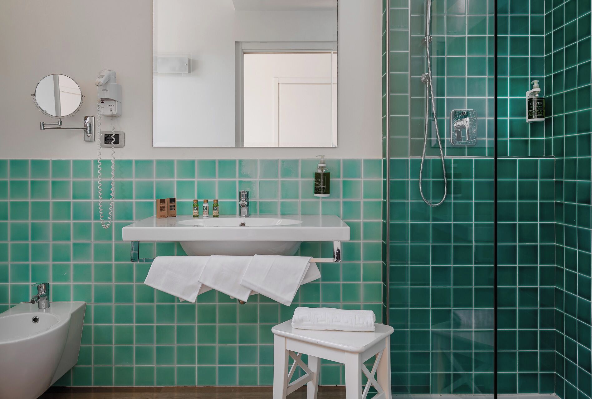 Un baño con un lavabo blanco, un espejo, un perchero para toallas, un cabezal de ducha de mano y paredes con azulejos verdes.
