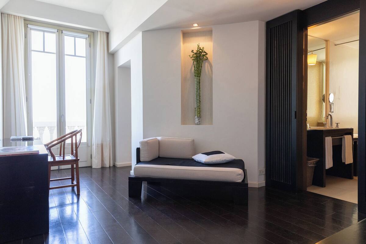 A modern hotel room with a dark wood floor, a sofa with cushions, a small table, a chair, a large window with curtains, a wall-mounted plant, and a bathroom visible through an open door.