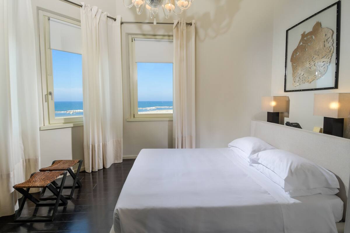 A hotel room with a bed, two windows with curtains, a view of the beach, and a chandelier.