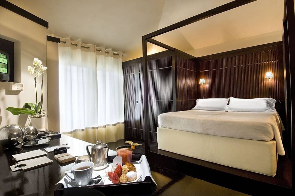 A hotel room with a bed, a desk with breakfast items, a plant, and a TV on the wall.