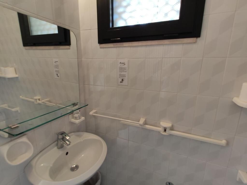 A bathroom with a glass shelf, a sink, a mirror, a handrail, and a window.