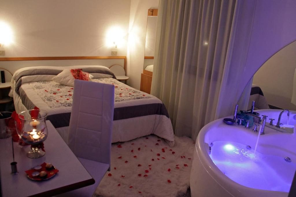 A hotel room with a bed, a bedside table with a candle, a large bathroom with a bathtub, and a mirror.