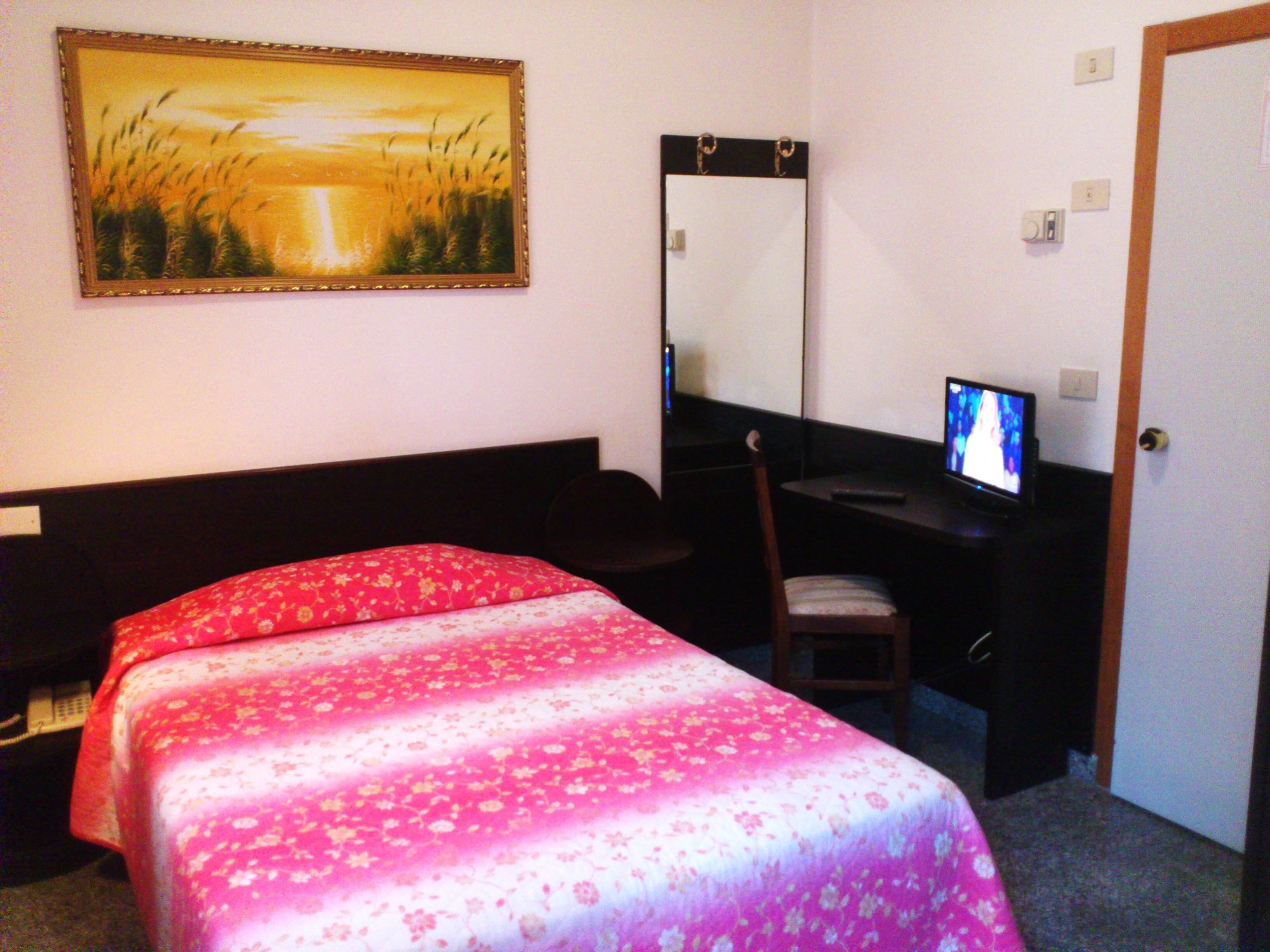Habitación de hotel con cama, escritorio con computadora, silla, espejo y un cuadro en la pared.
