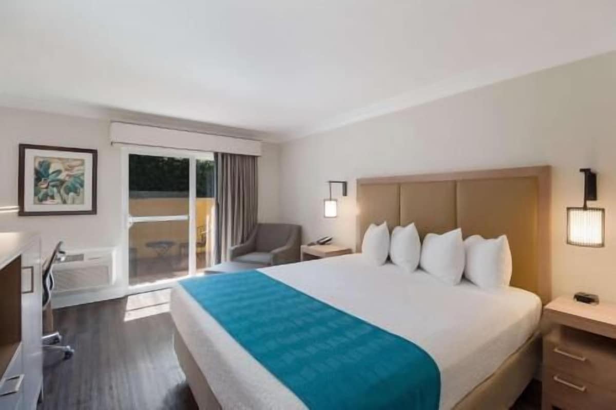 Una habitación de hotel con cama, escritorio, silla, mesita de noche y lámpara de pared.