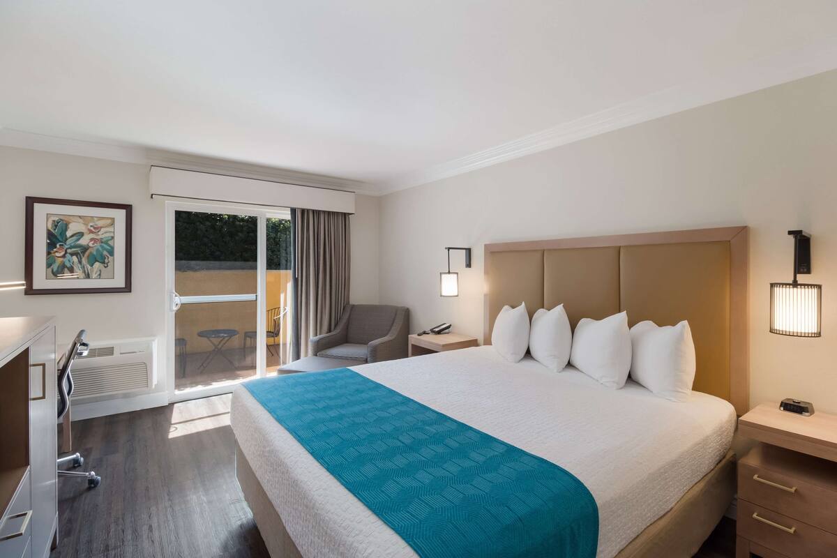 A hotel room with a large bed, a desk with a chair, a small table, a chair, a nightstand, and a wall-mounted lamp.