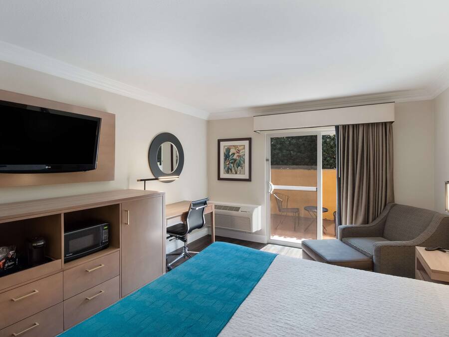 A hotel room with a bed, a desk with a chair, a television, a microwave, a large window with curtains, and a balcony with a table and chairs.