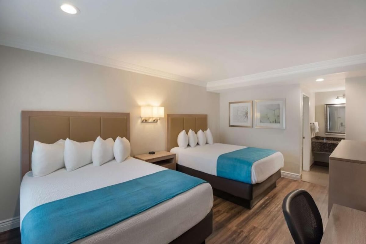 Habitación de hotel con dos camas, un escritorio y un baño visible a través de una puerta abierta.