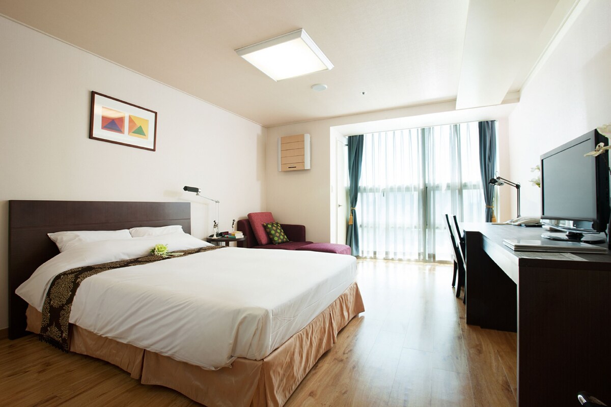 A hotel room with a large bed, a desk with a computer, a chair, a TV, and a window with curtains.