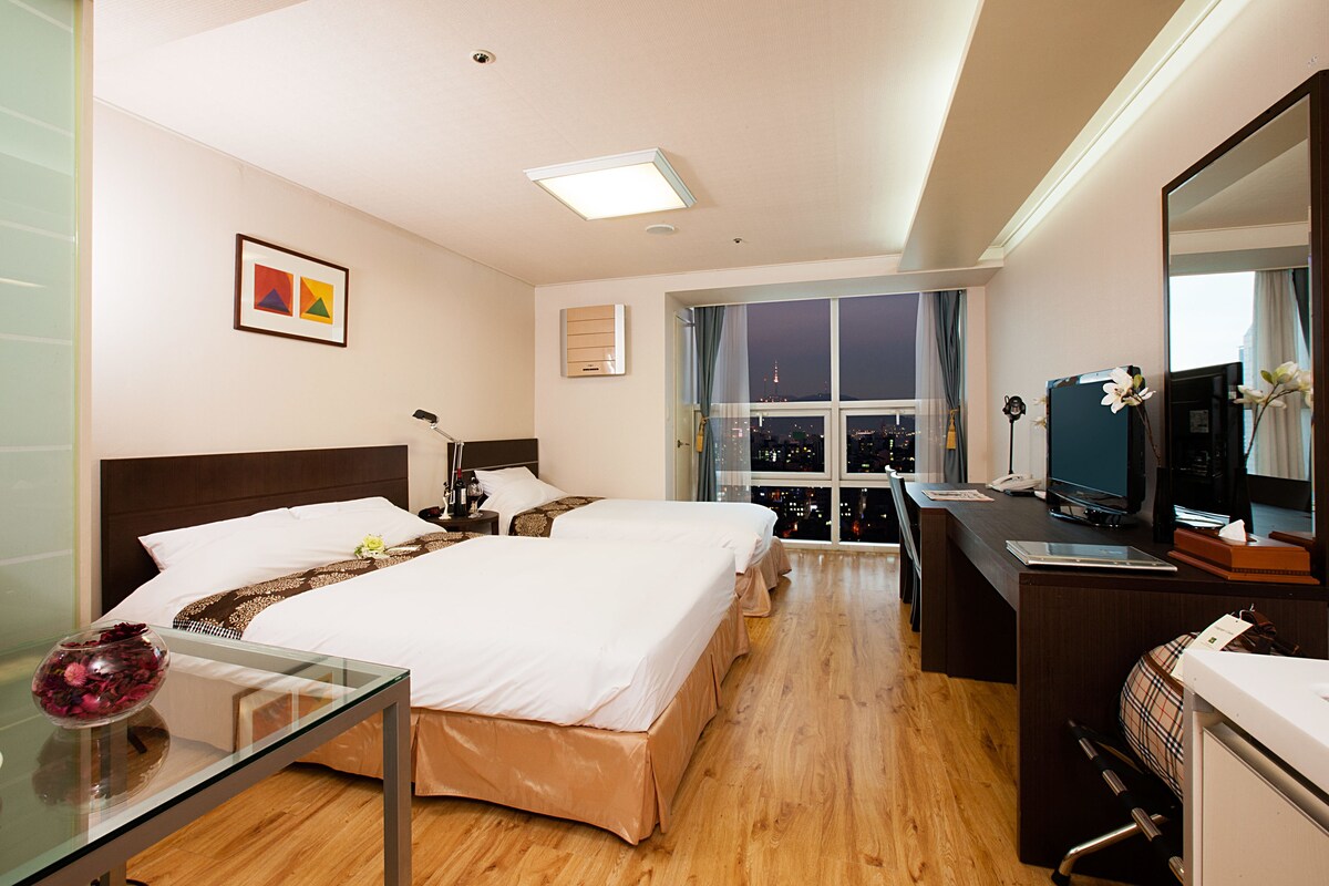 A hotel room with two beds, a desk with a computer, a mirror, and a window with a city view.