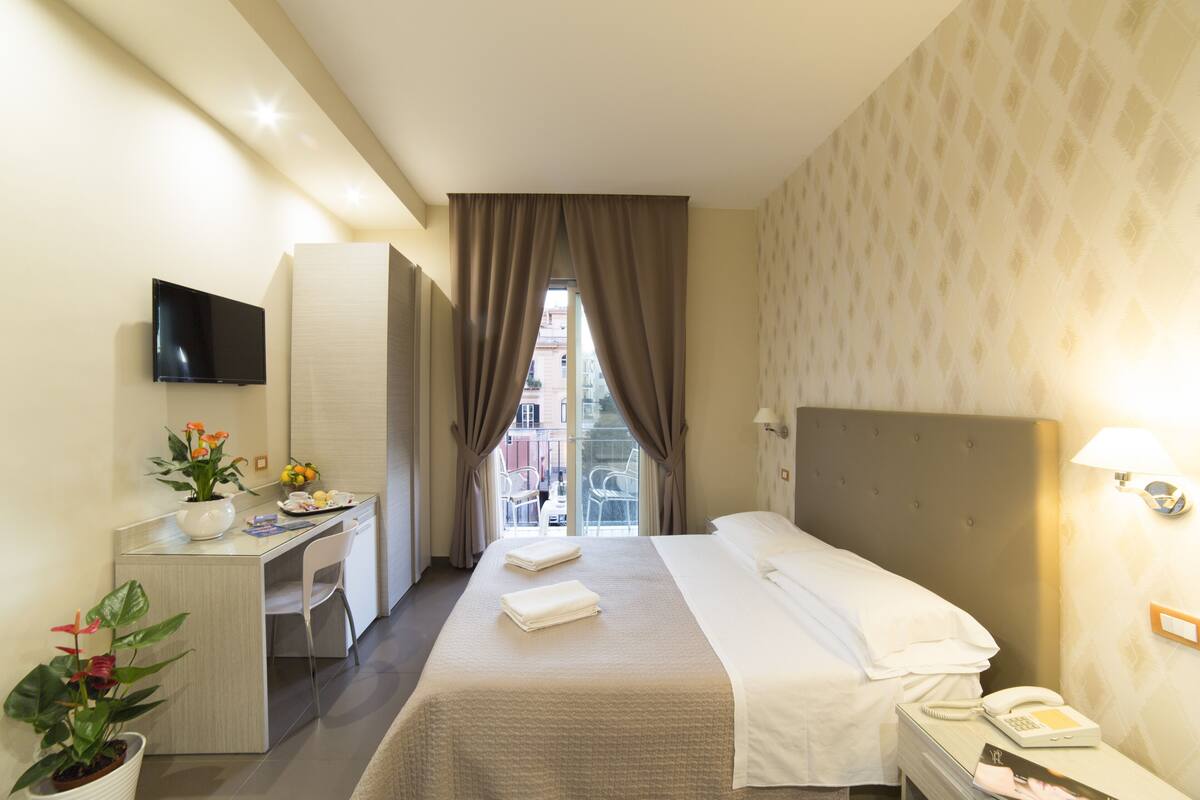 Ein Hotelzimmer mit Bett, Schreibtisch, Fernseher, Telefon und einem Fenster mit Vorhängen.
