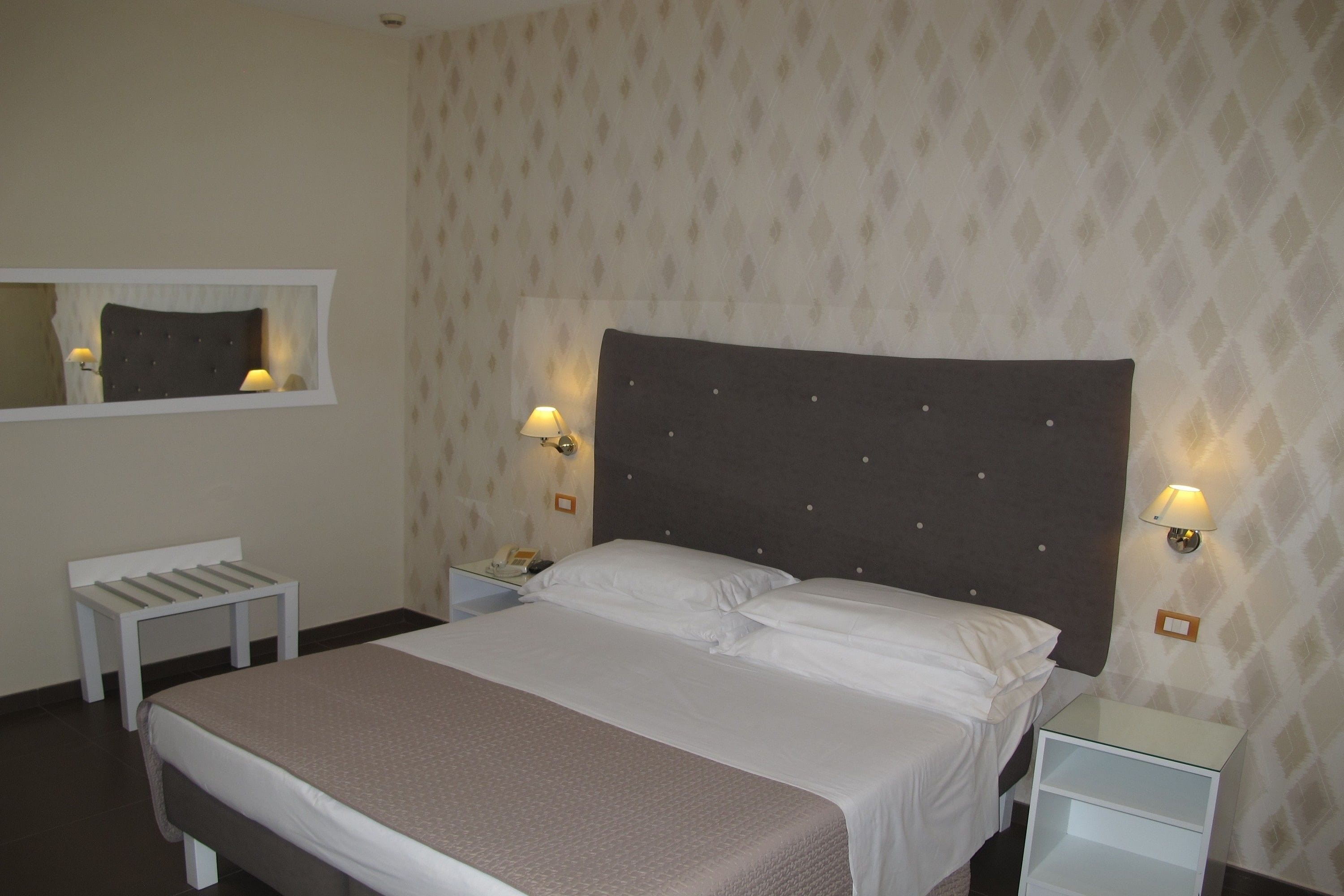 Quarto de hotel bem arrumado com uma cama grande, duas mesas de cabeceira com luminárias, um pequeno banco e um espelho sobre a cama.