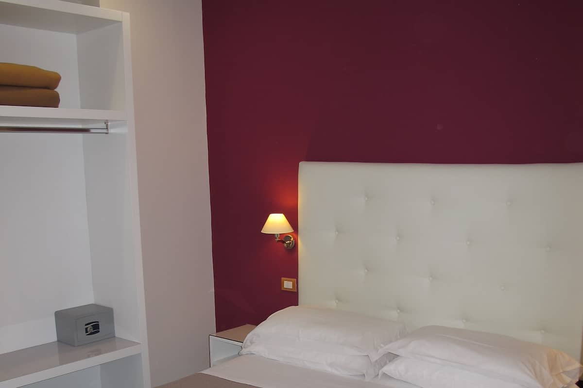 Ein ordentlich bezogenes Bett mit weißem Kopfteil, ein Nachttisch mit Lampe und ein Regal mit gefalteter Decke.