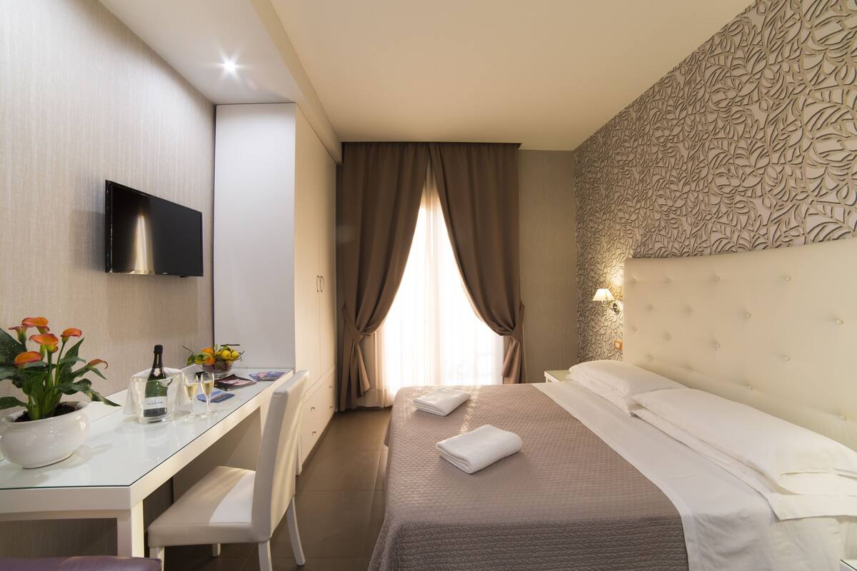Ein Hotelzimmer mit einem Bett, einem Schreibtisch mit Stuhl, einem an der Wand befestigten Fernseher und einem Fenster mit Vorhängen.