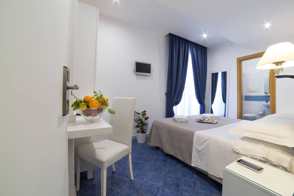 Ein Hotelzimmer mit Bett, Nachttisch, Stuhl, Schreibtisch mit Obstschale und einem Topf mit Pflanze sowie einem Fernseher.