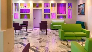 Un moderno vestíbulo con piso de mármol, sofás verdes, sillas púrpuras y estanterías iluminadas con objetos decorativos.