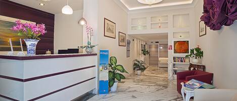 Zona de recepción de un hotel con suelo de mármol, mostrador de recepción, plantas en macetas y una araña.