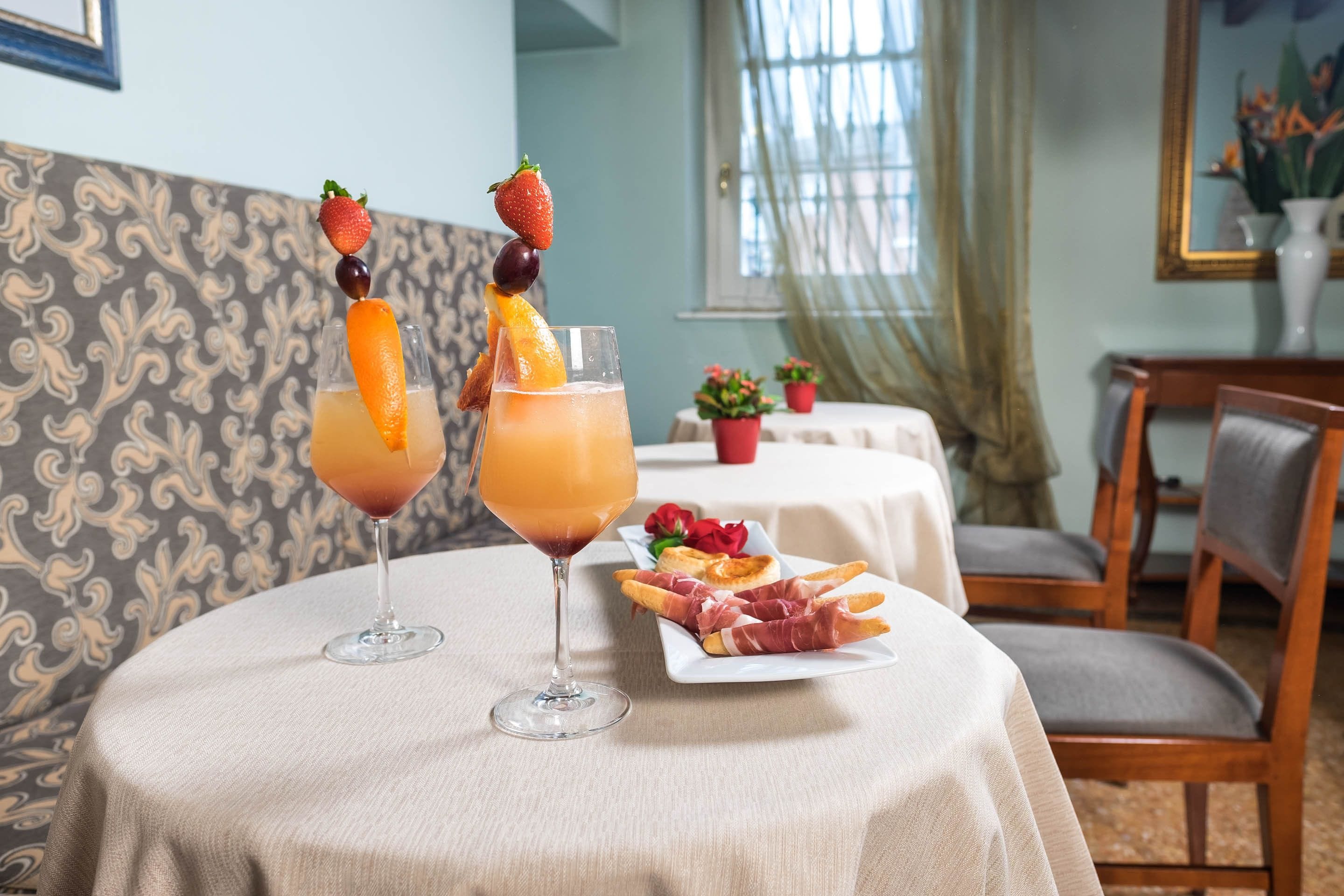 A dining table with two drinks garnished with fruits, a plate of prosciutto, and a floral centerpiece.