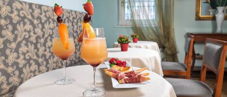 A dining table with two drinks garnished with fruits, a plate of prosciutto, and a floral centerpiece.