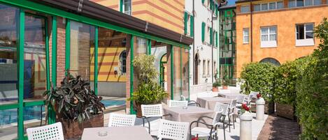 Un patio exterior con mesas y sillas, una piscina y edificios con contraventanas verdes y fachadas a rayas.