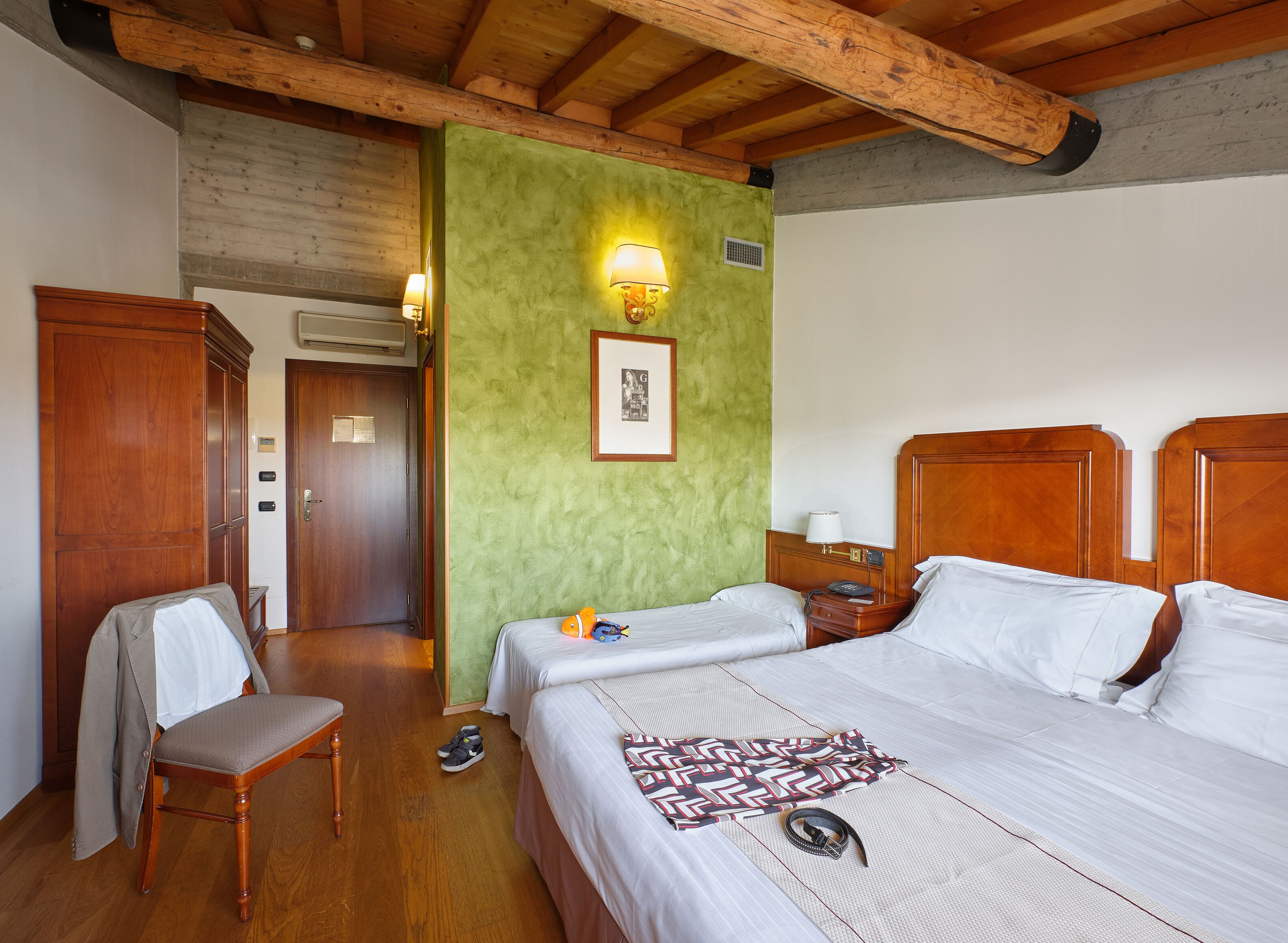 A hotel room with two beds, wooden headboards, a green accent wall, a framed picture, and a chair with a jacket.