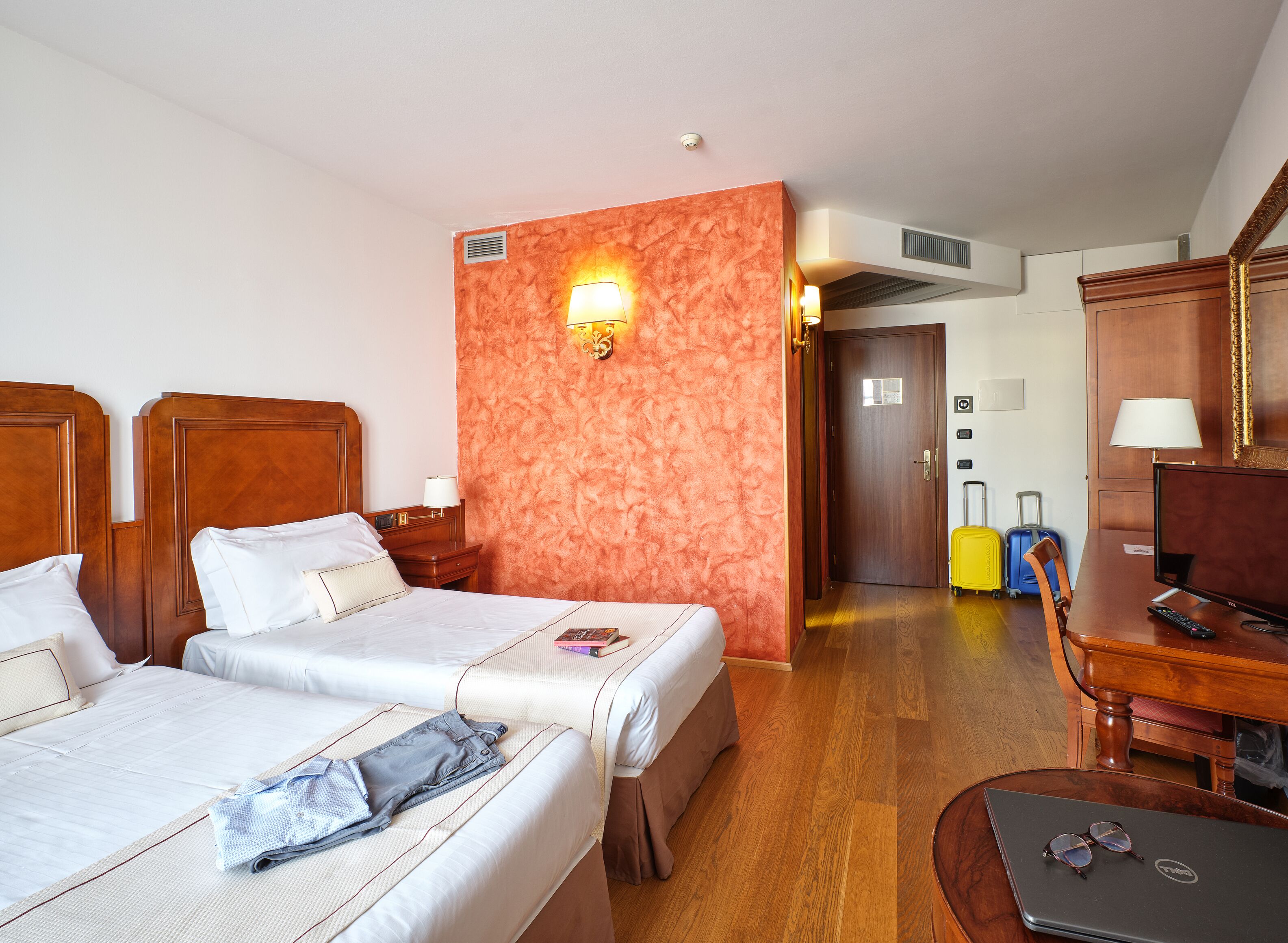 A hotel room with two beds, a desk with a computer, a TV, and a desk lamp. There is a suitcase and a chair in the corner.